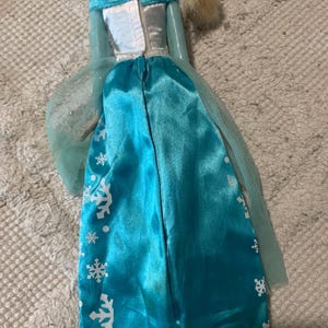 May include: A doll with blonde hair tied in a ponytail, wearing a long-sleeved dress with a white bodice and a shiny turquoise skirt featuring a white snowflake pattern. The dress includes sheer light blue cape panels attached at the shoulders.