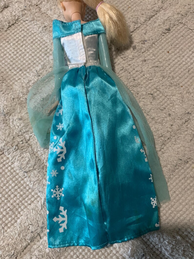 May include: A doll with blonde hair tied in a ponytail, wearing a long-sleeved dress with a white bodice and a shiny turquoise skirt featuring a white snowflake pattern. The dress includes sheer light blue cape panels attached at the shoulders.