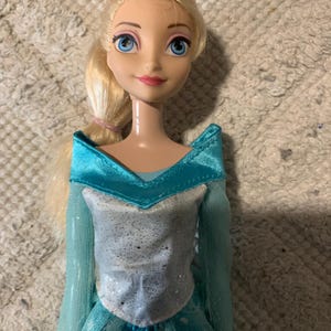 May include: A doll with blonde hair styled in a side ponytail, blue eyes, and pink lips. The doll wears a light blue dress featuring a silver glittery bodice, a wide collar, and long sheer sleeves, with a skirt patterned with white snowflake designs.