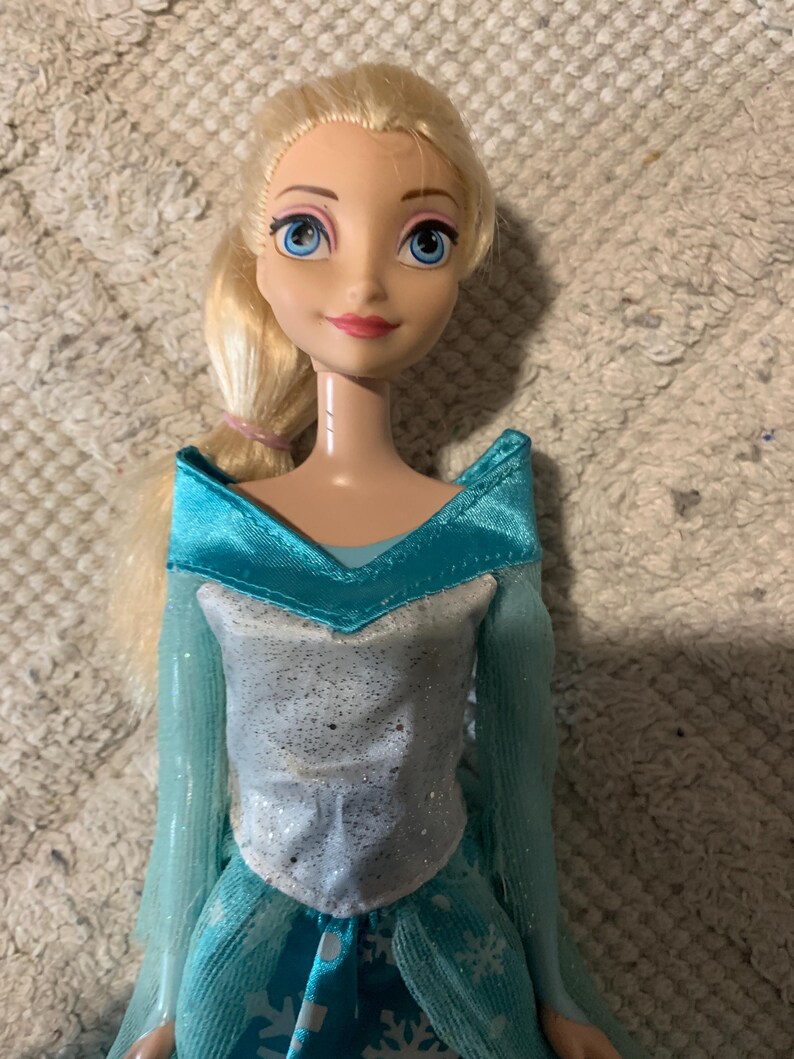 May include: A doll with blonde hair styled in a side ponytail, blue eyes, and pink lips. The doll wears a light blue dress featuring a silver glittery bodice, a wide collar, and long sheer sleeves, with a skirt patterned with white snowflake designs.