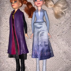 Puede incluir: Dos mu&ntilde;ecas que representan personajes de Frozen. La mu&ntilde;eca de la izquierda tiene el pelo rojo en una coleta, viste un abrigo morado sobre un vestido negro y botas negras. La mu&ntilde;eca de la derecha tiene el pelo rubio recogido en una coleta.