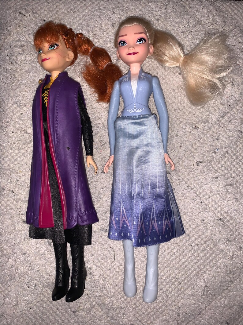 Puede incluir: Dos mu&ntilde;ecas que representan personajes de Frozen. La mu&ntilde;eca de la izquierda tiene el pelo rojo en una coleta, viste un abrigo morado sobre un vestido negro y botas negras. La mu&ntilde;eca de la derecha tiene el pelo rubio recogido en una coleta.