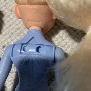 Puede incluir: Una mu&ntilde;eca con cabello rubio y un corpi&ntilde;o azul claro que presenta un patr&oacute;n de copo de nieve moldeado en la espalda. El cuello de la mu&ntilde;eca tiene grabado el texto Disney. La parte posterior del corpi&ntilde;o incluye un recorte rectangular.
