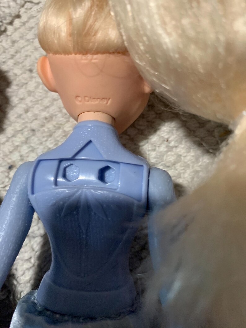 Puede incluir: Una mu&ntilde;eca con cabello rubio y un corpi&ntilde;o azul claro que presenta un patr&oacute;n de copo de nieve moldeado en la espalda. El cuello de la mu&ntilde;eca tiene grabado el texto Disney. La parte posterior del corpi&ntilde;o incluye un recorte rectangular.