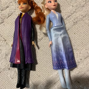 Puede incluir: Dos mu&ntilde;ecas que representan personajes de Frozen. La mu&ntilde;eca de la izquierda tiene el pelo casta&ntilde;o rojizo en una trenza, viste un abrigo morado sobre un vestido negro y botas negras. La mu&ntilde;eca de la derecha tiene el pelo rubio y viste un top azul.