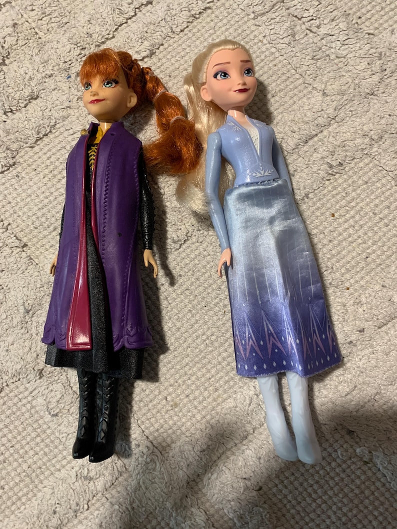 Puede incluir: Dos mu&ntilde;ecas que representan personajes de Frozen. La mu&ntilde;eca de la izquierda tiene el pelo casta&ntilde;o rojizo en una trenza, viste un abrigo morado sobre un vestido negro y botas negras. La mu&ntilde;eca de la derecha tiene el pelo rubio y viste un top azul.