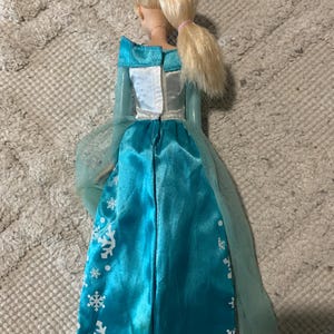 May include: A doll with blonde hair tied in a ponytail, wearing a long-sleeved dress with a white bodice and a bright blue satin skirt. The skirt features a white snowflake pattern and a sheer, light blue tulle cape attached at the shoulders.