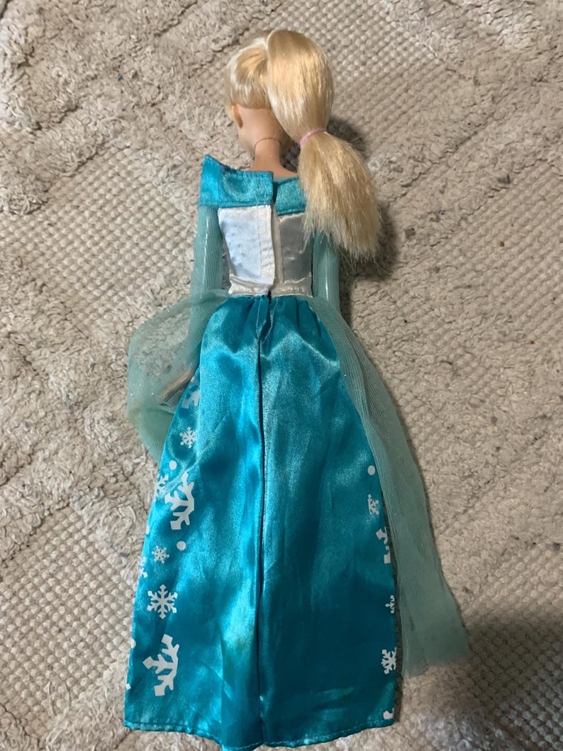 May include: A doll with blonde hair tied in a ponytail, wearing a long-sleeved dress with a white bodice and a bright blue satin skirt. The skirt features a white snowflake pattern and a sheer, light blue tulle cape attached at the shoulders.