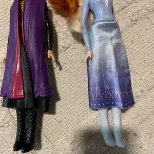 Puede incluir: Dos mu&ntilde;ecas una al lado de la otra. La mu&ntilde;eca de la izquierda tiene el pelo rojo, un abrigo morado sobre un conjunto negro y botas negras. La mu&ntilde;eca de la derecha tiene el pelo rubio, una blusa azul claro de manga larga, una falda azul degradada con patrones de copos de nieve blancos y botas blancas.