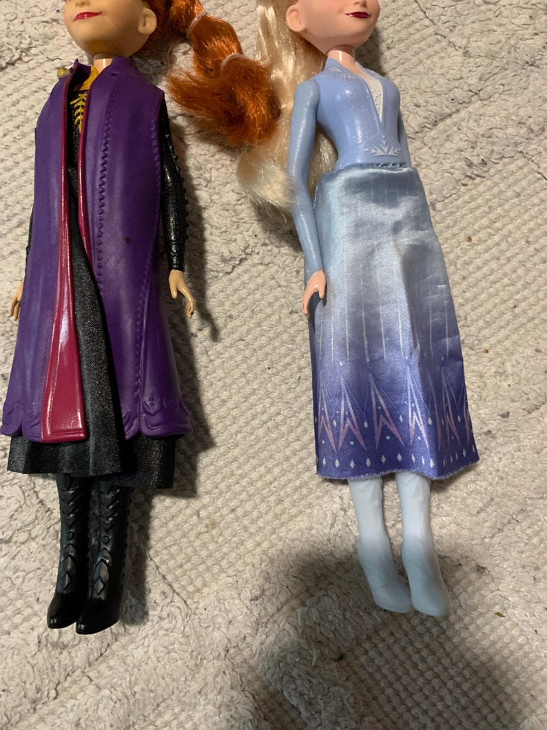 Puede incluir: Dos mu&ntilde;ecas una al lado de la otra. La mu&ntilde;eca de la izquierda tiene el pelo rojo, un abrigo morado sobre un conjunto negro y botas negras. La mu&ntilde;eca de la derecha tiene el pelo rubio, una blusa azul claro de manga larga, una falda azul degradada con patrones de copos de nieve blancos y botas blancas.