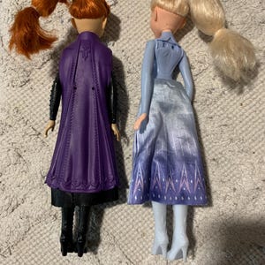 Puede incluir: Dos mu&ntilde;ecas de pie una al lado de la otra. La mu&ntilde;eca de la izquierda tiene el pelo casta&ntilde;o rojizo en una coleta, una capa morada moldeada, mangas negras y botas negras. La mu&ntilde;eca de la derecha tiene el pelo rubio en una coleta y falda azul.