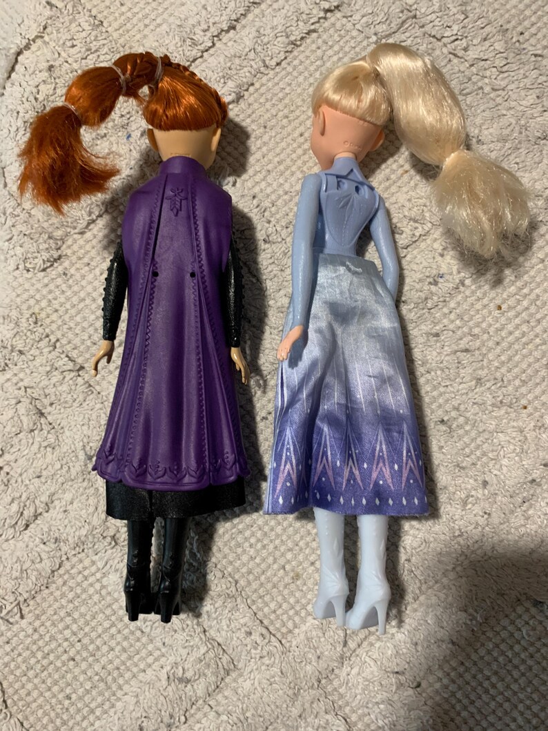 Puede incluir: Dos mu&ntilde;ecas de pie una al lado de la otra. La mu&ntilde;eca de la izquierda tiene el pelo casta&ntilde;o rojizo en una coleta, una capa morada moldeada, mangas negras y botas negras. La mu&ntilde;eca de la derecha tiene el pelo rubio en una coleta y falda azul.