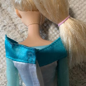 May include: A doll with blonde hair tied in a ponytail with a pink elastic band. The doll wears a teal and white dress with an off-the-shoulder neckline. The word DISNEY is embossed on the back of the doll's neck. The dress has a white back panel.