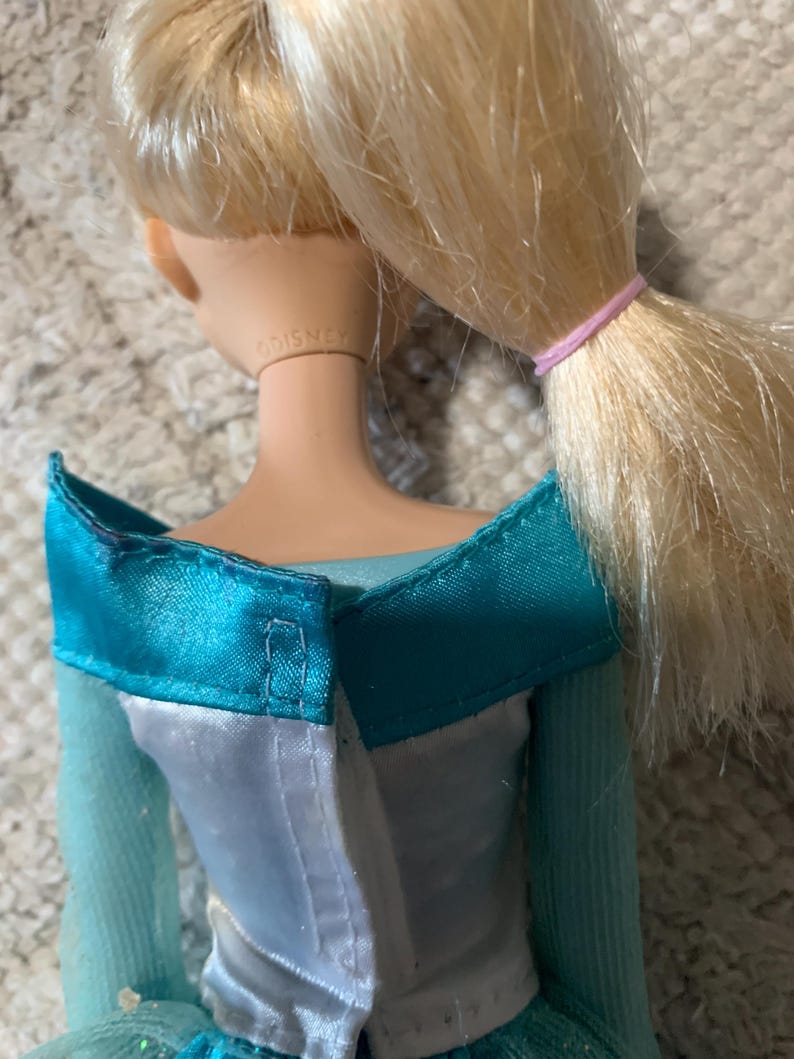 May include: A doll with blonde hair tied in a ponytail with a pink elastic band. The doll wears a teal and white dress with an off-the-shoulder neckline. The word DISNEY is embossed on the back of the doll's neck. The dress has a white back panel.