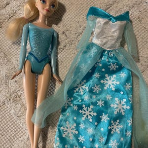 May include: A blonde doll with blue eyes wearing a shimmering light blue leotard, accompanied by a separate floor-length gown featuring a silver bodice, sheer light blue sleeves, and a bright blue skirt decorated with a white snowflake pattern.