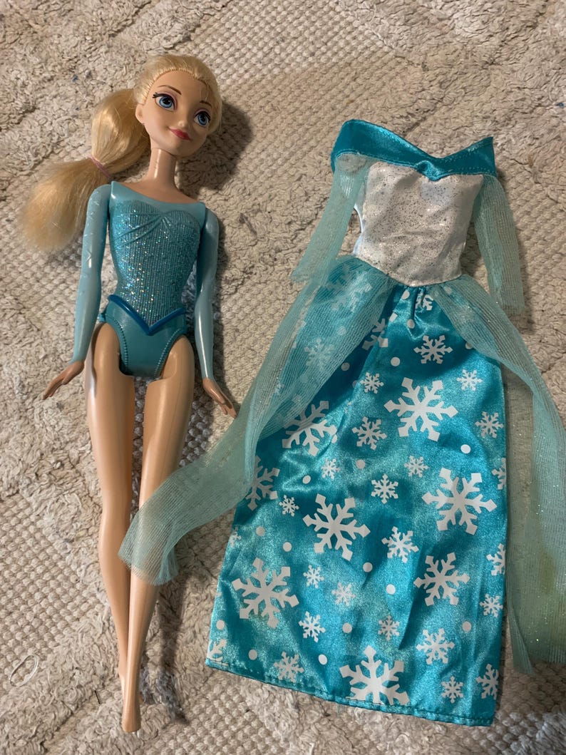 May include: A blonde doll with blue eyes wearing a shimmering light blue leotard, accompanied by a separate floor-length gown featuring a silver bodice, sheer light blue sleeves, and a bright blue skirt decorated with a white snowflake pattern.