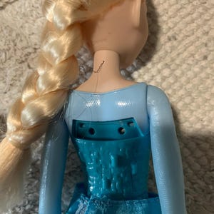 May include: A doll with blonde hair styled in a long side braid, wearing a light blue bodice with a textured, geometric pattern on the back. The back of the bodice features an open rectangular cutout with two visible screw holes and a fabric skirt.