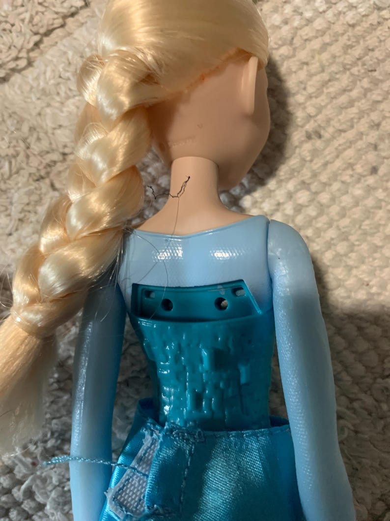 May include: A doll with blonde hair styled in a long side braid, wearing a light blue bodice with a textured, geometric pattern on the back. The back of the bodice features an open rectangular cutout with two visible screw holes and a fabric skirt.