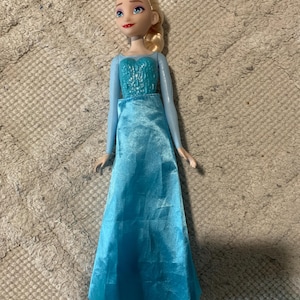 May include: A doll with blonde hair styled in a side braid, featuring blue eyes and red lips. The doll wears a light blue long-sleeved gown with a textured bodice and a smooth, floor-length satin skirt that extends down to the feet.