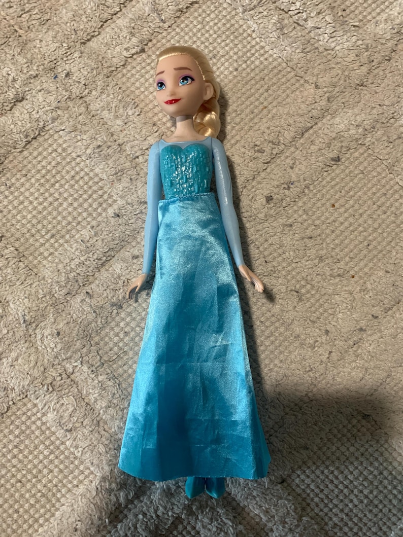 May include: A doll with blonde hair styled in a side braid, featuring blue eyes and red lips. The doll wears a light blue long-sleeved gown with a textured bodice and a smooth, floor-length satin skirt that extends down to the feet.