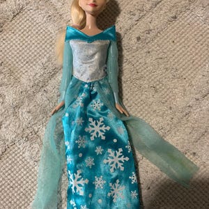 May include: A doll with blonde hair styled in a side ponytail, featuring blue eyes and pink lips. The doll wears a long-sleeved dress with a silver bodice and a bright blue skirt adorned with white snowflake patterns and sheer blue fabric accents.