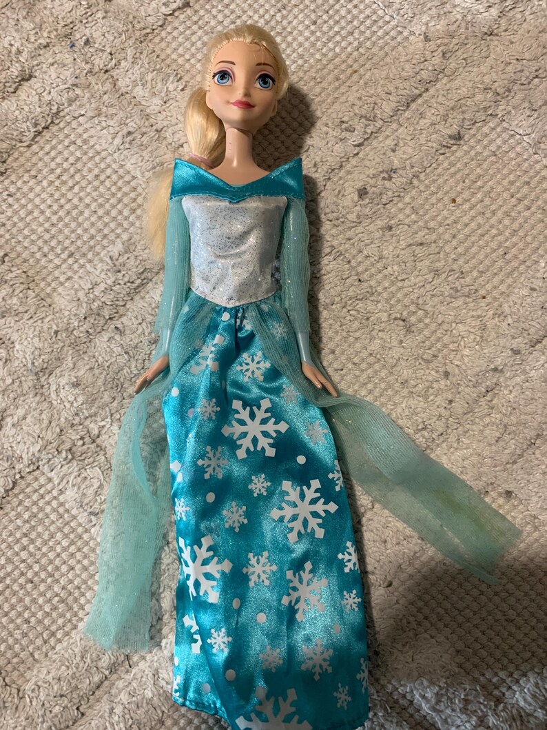May include: A doll with blonde hair styled in a side ponytail, featuring blue eyes and pink lips. The doll wears a long-sleeved dress with a silver bodice and a bright blue skirt adorned with white snowflake patterns and sheer blue fabric accents.