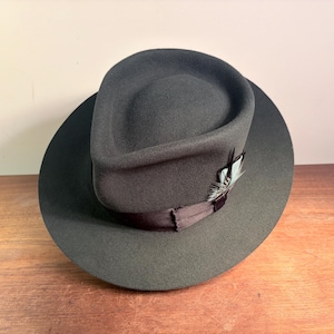 Può includere: Un cappello fedora in feltro grigio scuro con tesa larga e corona pizzicata. Un nastro in raso marrone scuro circonda la base della corona, arricchito da un piccolo dettaglio in piuma decorativa inserito sul lato del nastro del cappello.