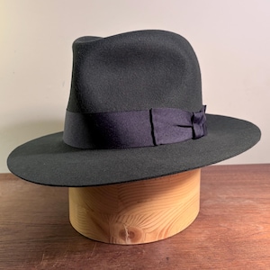 May include: A dark charcoal gray fedora hat featuring a structured crown with a center crease and a wide brim. The hat is accented with a dark navy blue grosgrain ribbon band that wraps around the base of the crown, finished with a small bow detail.