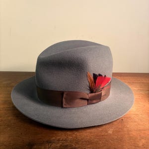 Peut inclure: Un chapeau fedora gris clair doté d'un large ruban en gros-grain marron autour de la calotte. Le chapeau est agrémenté d'un groupe de petites plumes aux couleurs vives rouge, orange et marron foncé insérées dans le ruban.