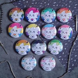 Può includere: Una collezione di dodici spille rotonde raffiguranti una creatura bianca dei cartoni animati con grandi occhi che tiene in mano un piccolo cuore. Ogni spilla mostra un diverso sfondo con bandiera arcobaleno, finitura glitter e dettagli a rombo.
