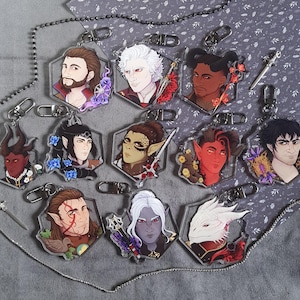 Op de afbeelding: Een collectie van elf zeshoekige acryl sleutelhangers met illustraties van diverse fantasy-personages. Elke bedel is voorzien van een zilverkleurige karabijnsluiting en toont unieke portretten met gevarieerde huidtinten, haarkleuren en hoorns.