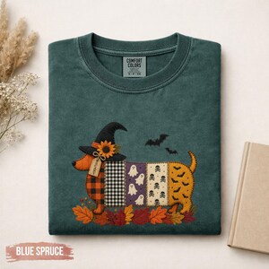 Peut inclure: Un t-shirt à col rond vert foncé avec un motif de teckel portant un chapeau de sorcière. Le corps du chien est composé de panneaux verticaux à motifs écossais, vichy, fantômes, crânes et chauves-souris, posé sur des feuilles d'automne.