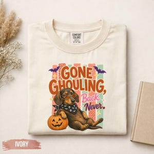 Può includere: Una t-shirt color avorio con la grafica di un bassotto che indossa occhiali da sole rotondi e una bandana con teschi, seduto accanto a una zucca. Il testo recita GONE GHOULING, Back Never, su strisce verticali colorate e pipistrelli.