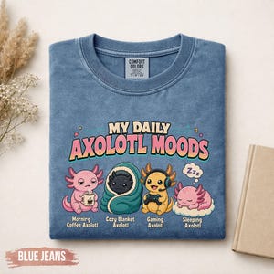 May include: A blue crewneck t-shirt featuring the text "MY DAILY AXOLOTL MOODS" and four axolotl illustrations labeled "Morning Coffee Axolotl," "Cozy Blanket Axolotl," "Gaming Axolotl," and "Sleeping Axolotl" with a Comfort Colors brand tag.
