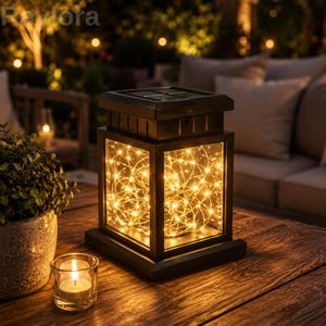 May include: A square black solar lantern with clear glass panels containing a cluster of warm white fairy lights. The top features a solar panel, and the brand name Raylora is visible in the upper left corner of the image.