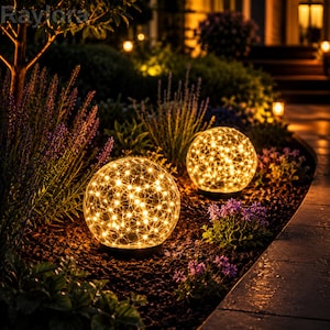 Puede incluir: Dos lámparas de jardín esféricas con textura de vidrio agrietado, cada una con una cadena de luces LED de color blanco cálido en su interior. Los globos están colocados sobre mantillo oscuro entre lavanda morada y follaje verde.