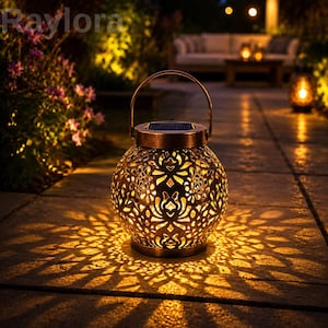 May include: A round, bronze-colored metal solar lantern with a handle and intricate cut-out patterns that cast a glowing, floral-like light projection onto the ground. The top features a small solar panel, and the word Raylora is visible.