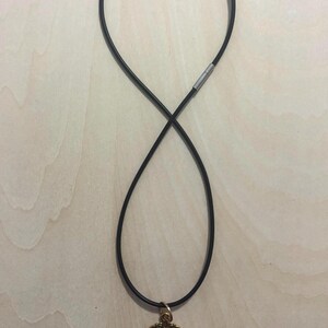 May include: A black cord necklace featuring a silver-toned metal clasp and an oval-shaped pendant. The pendant has an ornate, antique-gold-colored metal frame with a textured, dark, and mottled pattern visible under a smooth, rounded glass surface.