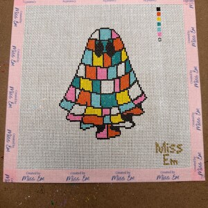 May include: A square needlepoint canvas featuring a colorful, patchwork-style ghost design in shades of blue, orange, pink, and yellow. The border is printed with the text "Created by Miss Em" repeatedly, and the logo "Miss Em" appears in the corner.