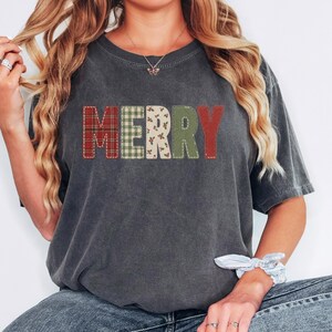 May include: A charcoal grey short-sleeve t-shirt featuring the word MERRY printed across the chest. Each letter has a distinct pattern: red plaid, green gingham, cream with holly berries, solid green, and solid red, all with a stitched border effect.