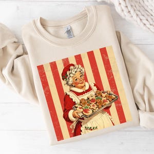 May include: A beige crewneck sweatshirt featuring a graphic print of a woman with white hair wearing a red dress and white apron, holding a metal tray of gingerbread cookies against a background of vertical red and cream stripes.