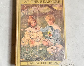 以下が含まれることがあります： ローラ・リー・ホープ著『The Bobbsey Twins at the Seashore』のヴィンテージハードカバー本。黄色の表紙には、花が咲く草地に座る二人の子供のカラーイラストが描かれています。テキストは黄色の縁取りに黒で印刷されています。