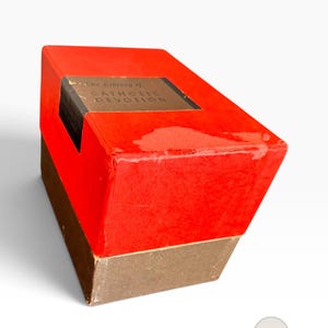May include: A rectangular box with a bright red lid and a dark brown base. The lid features a black rectangular label with the gold text The Library of CATHOLIC DEVOTION. The box shows signs of wear on the red surface and edges of the lid.