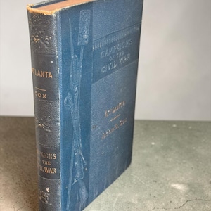 May include: A blue hardcover book titled Campaigns of the Civil War: Atlanta by Cox. The front cover features a debossed illustration of a sword and flag, while the spine displays the title, author, and publisher in gold lettering.