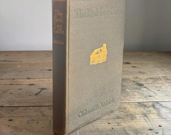 以下が含まれることがあります： チルソン・D・オルドリッチ著『The Real Log Cabin』のハードカバー本。質感のあるオリーブグリーンの表紙で、前面には小さな金色の丸太小屋のイラストが描かれ、背表紙には金色の文字でタイトルと著者名が記されています。