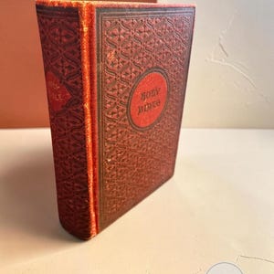 May include: A vintage hardcover book with a deep red fabric cover featuring an intricate embossed geometric pattern. The front cover displays a central red circular medallion with the text Holy Bible printed in dark, weathered lettering.