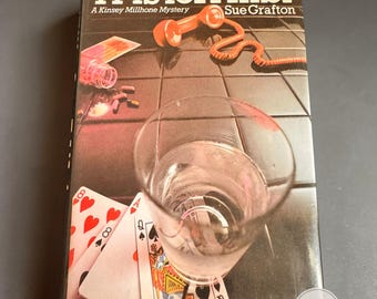 以下が含まれることがあります： スー・グラフトン著のペーパーバック「A is for Alibi」。表紙には黒いタイル張りの表面が描かれており、オレンジ色の電話の受話器、透明な水の入ったグラス、散らばったトランプ、そして錠剤の入った小さな瓶が配置されています。