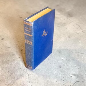 May include: A blue hardcover book titled The Blind Goddess by Arthur Train. The spine features gold lettering and decorative lines. The front cover displays a small gold emblem of an open book. The pages have a light tan, aged appearance.