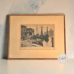 May include: A framed black and white etching depicting a harbor scene with several docked boats, a small wooden pier, and a building. The artwork is set within a light-colored mat and a thin wooden frame, featuring handwritten text at the bottom.