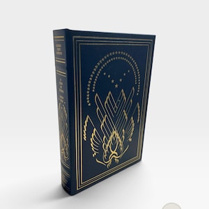 May include: A dark blue hardcover book with gold foil stamping on the front cover and spine. The front features a stylized wing design and an arch of stars. The spine text reads Signed First Edition, A God in Ruins, and Leon Uris.